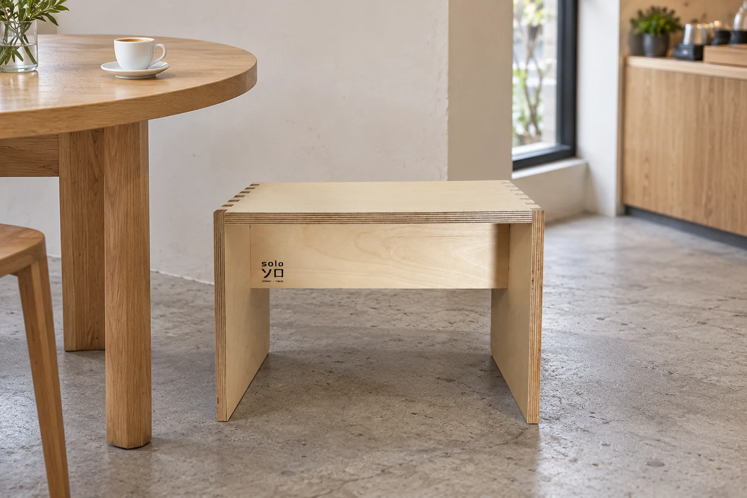 The Kumi plywood stool beside a pale oak table in a quiet contemporary cafe, lit by a window.